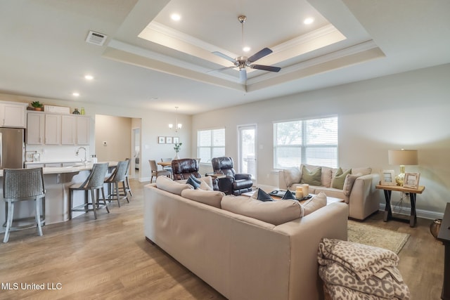 living room featuring ceiling fan with notable chandelier, sink, a tray ceiling, crown molding, and light hardwood / wood-style flooring