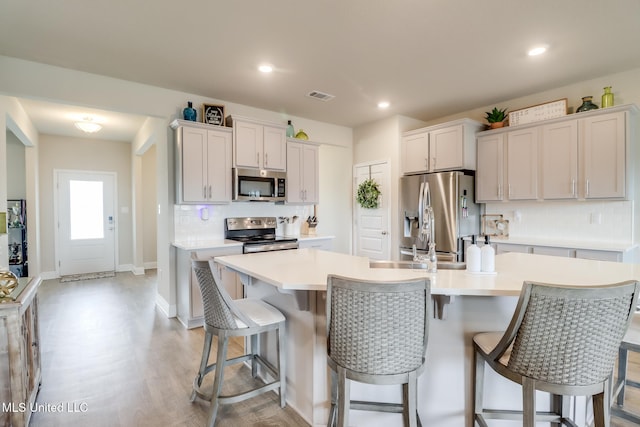 kitchen with a breakfast bar area, appliances with stainless steel finishes, a center island with sink, decorative backsplash, and light wood-type flooring