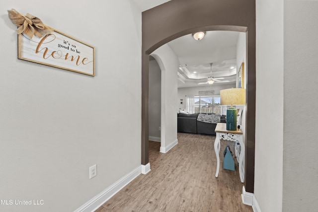 corridor featuring baseboards, arched walkways, a raised ceiling, and wood finished floors