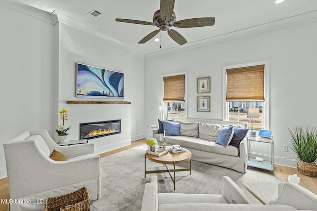 living area with visible vents, crown molding, ceiling fan, wood finished floors, and a glass covered fireplace