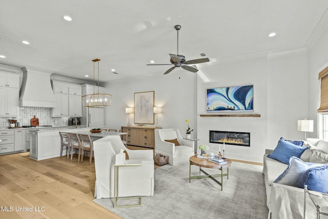 living room featuring crown molding, recessed lighting, light wood-style flooring, ceiling fan with notable chandelier, and a glass covered fireplace