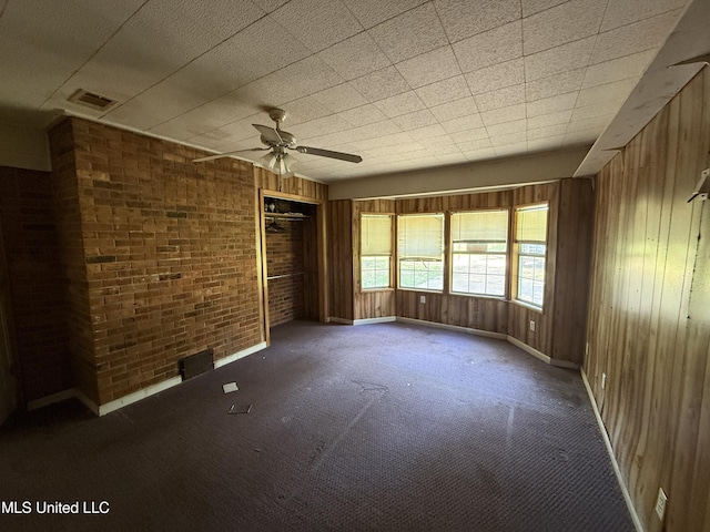 carpeted empty room with a ceiling fan, baseboards, wooden walls, and brick wall