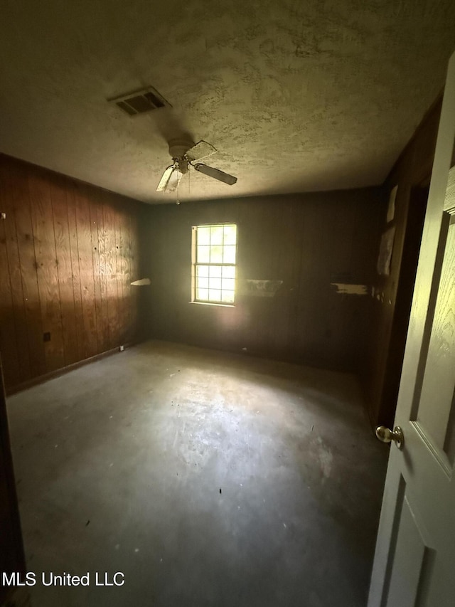 spare room featuring unfinished concrete flooring, a ceiling fan, wooden walls, and a textured ceiling