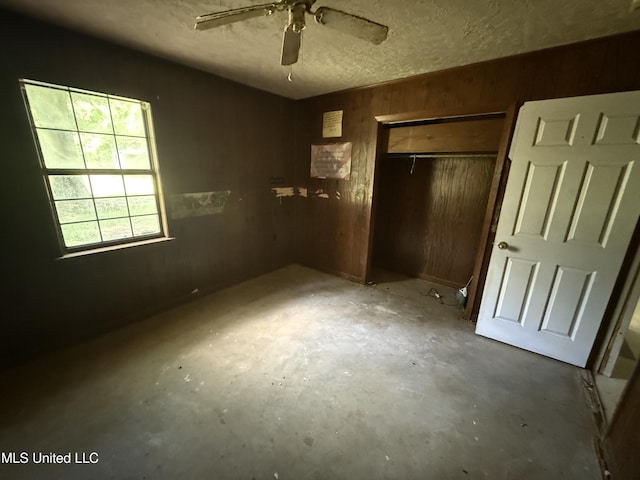 unfurnished bedroom featuring wood walls, unfinished concrete flooring, a closet, a textured ceiling, and ceiling fan