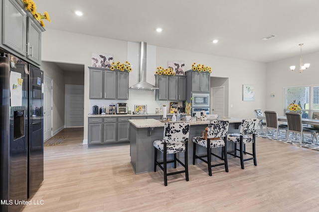 kitchen with stainless steel microwave, an island with sink, black fridge, wall chimney exhaust hood, and gray cabinets