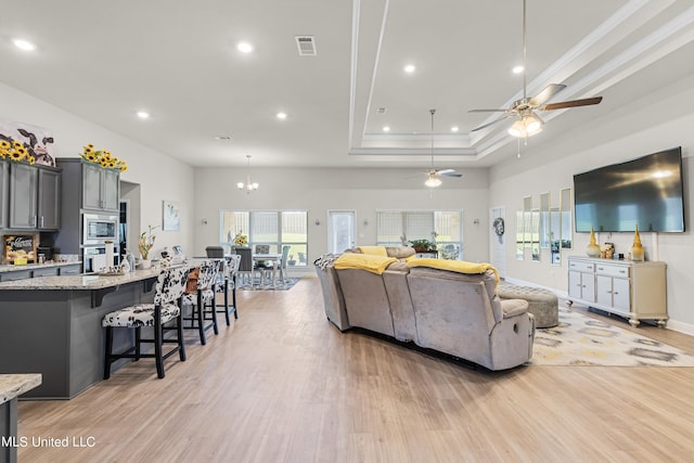 living room with ceiling fan with notable chandelier, light wood-type flooring, and a raised ceiling