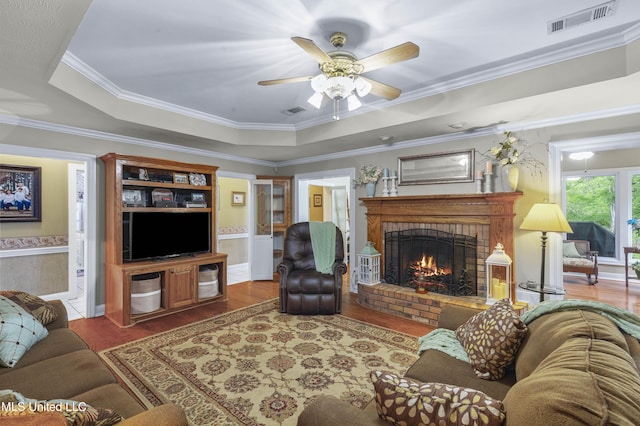 living area featuring wood finished floors, a tray ceiling, a brick fireplace, a ceiling fan, and ornamental molding