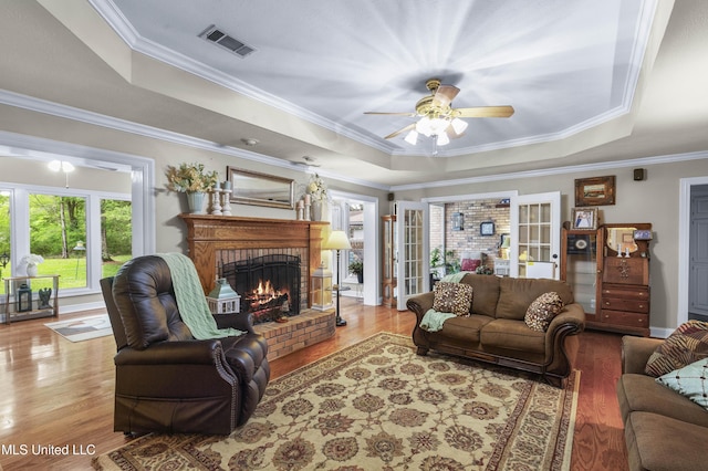 living room with a raised ceiling, ceiling fan, wood finished floors, crown molding, and a brick fireplace