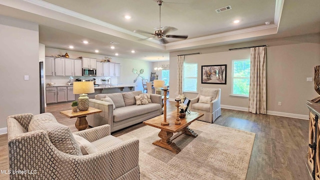 living room with wood-type flooring, ceiling fan with notable chandelier, crown molding, and a tray ceiling