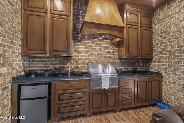 kitchen with custom exhaust hood, stainless steel refrigerator, light hardwood / wood-style flooring, sink, and brick wall