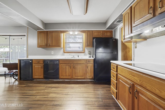kitchen with light countertops, under cabinet range hood, brown cabinets, a sink, and black appliances