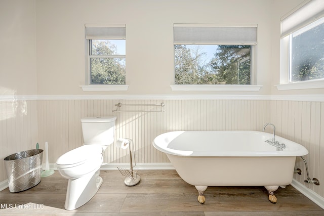 bathroom with wood-type flooring, a tub to relax in, toilet, and a healthy amount of sunlight