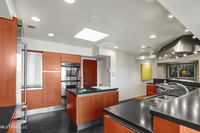 kitchen with double oven, wall chimney exhaust hood, an island with sink, sink, and a skylight