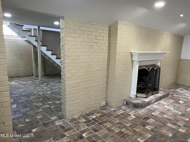 finished below grade area featuring a fireplace with raised hearth, brick floor, recessed lighting, brick wall, and stairs