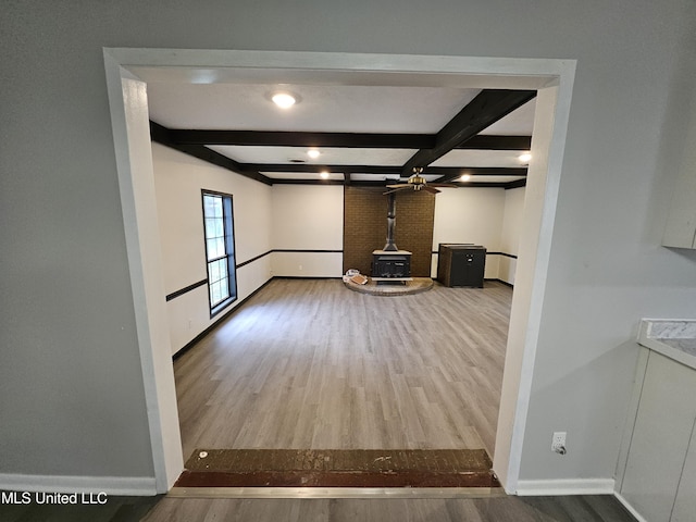 unfurnished living room featuring a ceiling fan, a wood stove, wood finished floors, baseboards, and beamed ceiling