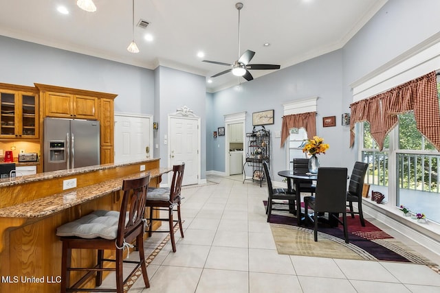 kitchen with a kitchen breakfast bar, stainless steel refrigerator with ice dispenser, ceiling fan, a towering ceiling, and light stone counters