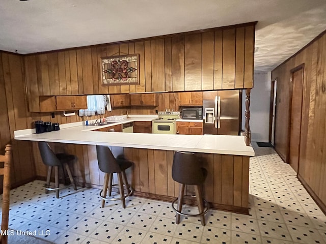 kitchen with a kitchen bar, kitchen peninsula, white electric stove, stainless steel fridge with ice dispenser, and wood walls