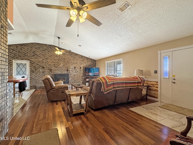 living room featuring ceiling fan, vaulted ceiling, a fireplace, hardwood / wood-style floors, and brick wall