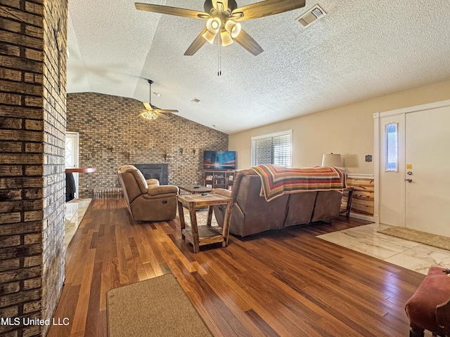 living room with a brick fireplace, ceiling fan, brick wall, wood-type flooring, and vaulted ceiling