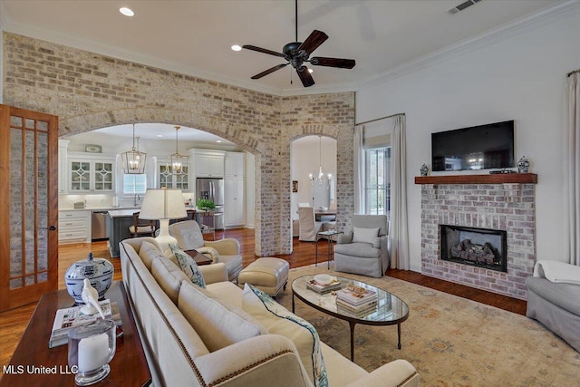 living room featuring light wood finished floors, a brick fireplace, crown molding, ceiling fan with notable chandelier, and arched walkways