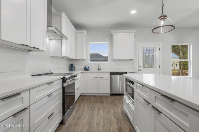 kitchen with light wood-type flooring, appliances with stainless steel finishes, white cabinets, pendant lighting, and wall chimney range hood