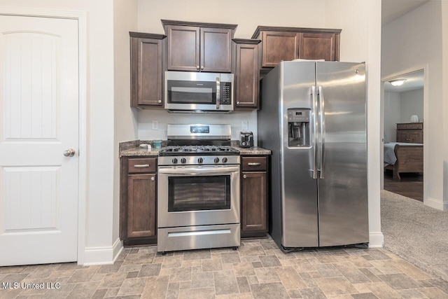 kitchen with stainless steel appliances, dark brown cabinets, light carpet, and dark stone counters