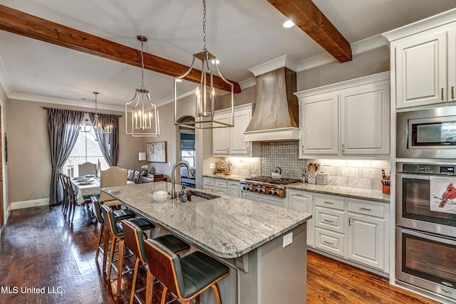 kitchen featuring premium range hood, stainless steel appliances, a kitchen island with sink, beam ceiling, and white cabinets