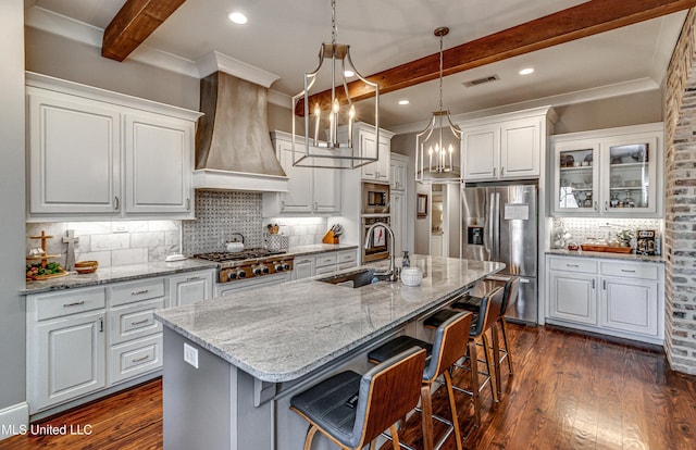 kitchen featuring premium range hood, beamed ceiling, white cabinetry, appliances with stainless steel finishes, and an island with sink