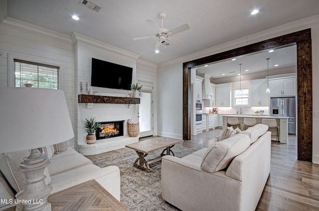 living area with crown molding, light wood-type flooring, a brick fireplace, a ceiling fan, and baseboards