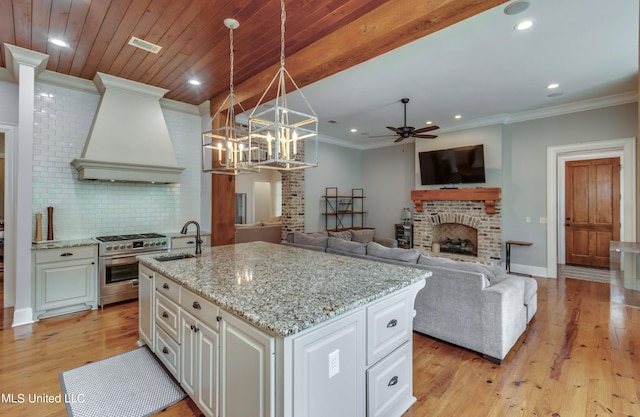 kitchen featuring custom range hood, high end range, a center island with sink, decorative light fixtures, and light wood-type flooring