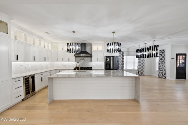 kitchen featuring white cabinets, black refrigerator with ice dispenser, wall chimney range hood, tasteful backsplash, and a center island with sink