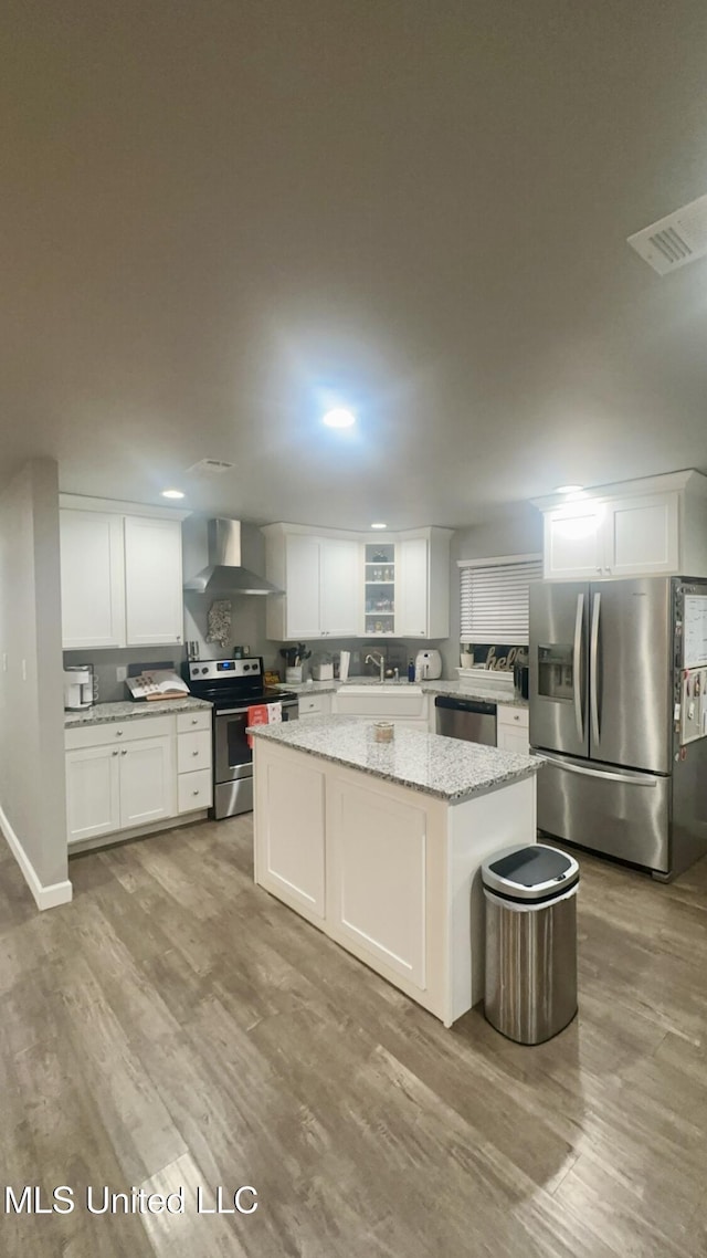 kitchen featuring a center island, wall chimney range hood, light stone counters, white cabinets, and appliances with stainless steel finishes