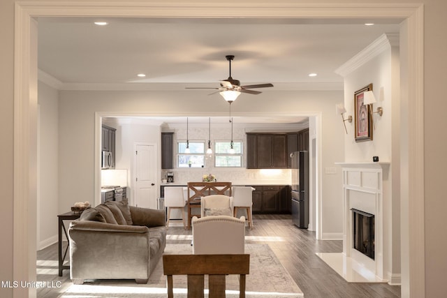 living area with ceiling fan, a fireplace with flush hearth, wood finished floors, baseboards, and ornamental molding