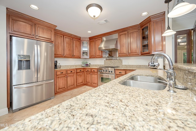 kitchen with appliances with stainless steel finishes, wall chimney exhaust hood, a sink, brown cabinets, and glass insert cabinets