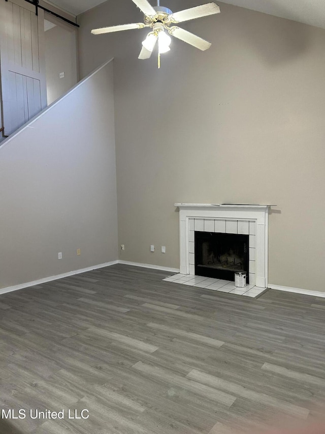 unfurnished living room with high vaulted ceiling, a tile fireplace, hardwood / wood-style flooring, ceiling fan, and a barn door