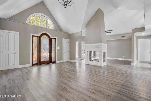 unfurnished living room with ceiling fan with notable chandelier, light hardwood / wood-style flooring, high vaulted ceiling, and a brick fireplace