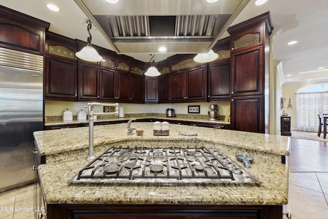 kitchen featuring a kitchen island with sink, light stone counters, stainless steel appliances, dark brown cabinets, and wall chimney exhaust hood