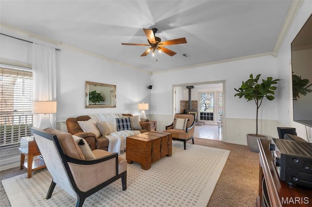 living area featuring ceiling fan, crown molding, wainscoting, light carpet, and a wood stove