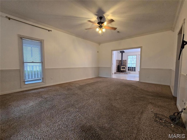 carpeted spare room with crown molding, wainscoting, ceiling fan, a wood stove, and a textured ceiling