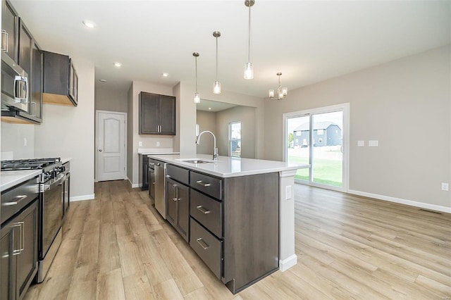 kitchen with stainless steel appliances, a sink, light wood finished floors, a chandelier, and a kitchen island with sink