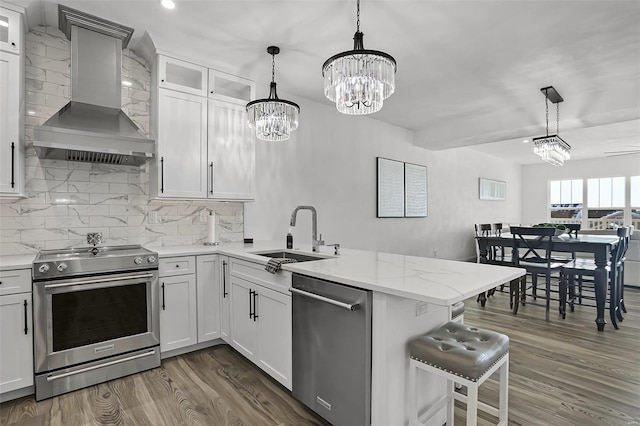 kitchen featuring appliances with stainless steel finishes, a sink, wall chimney range hood, a chandelier, and a peninsula