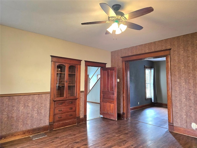 empty room featuring wallpapered walls, wainscoting, ceiling fan, dark wood-style floors, and baseboards