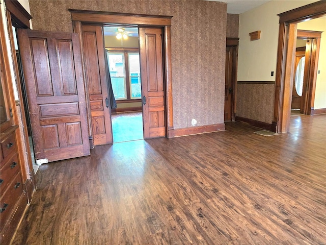 unfurnished room featuring dark wood finished floors, baseboards, a ceiling fan, wallpapered walls, and a wainscoted wall