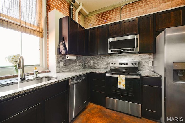kitchen with stainless steel appliances, healthy amount of natural light, light stone countertops, a sink, and brick wall