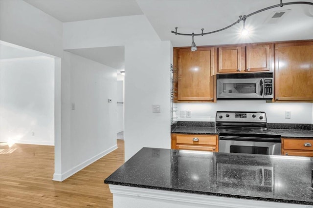 kitchen featuring stainless steel appliances, light wood-type flooring, brown cabinetry, rail lighting, and dark stone counters