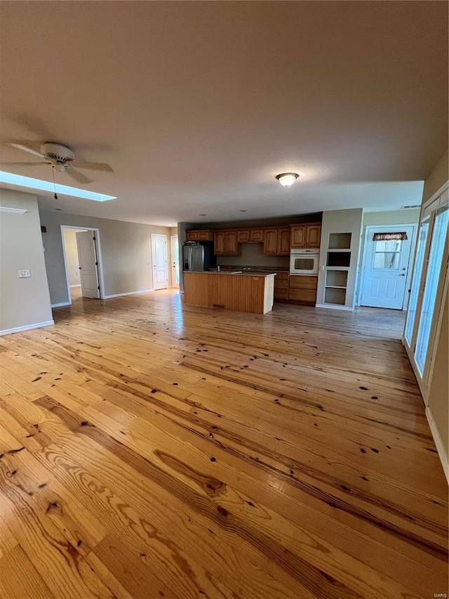 unfurnished living room featuring baseboards, light wood-type flooring, a ceiling fan, and a skylight