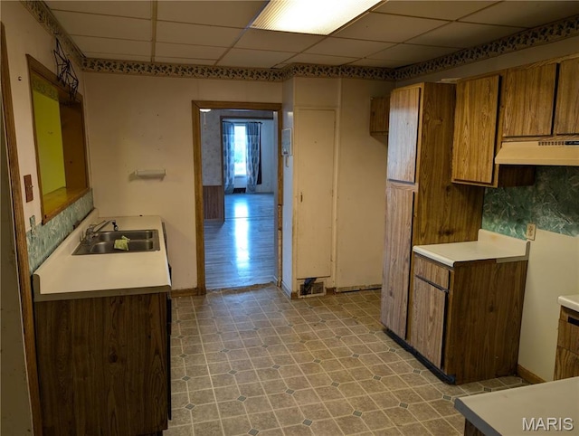 kitchen with a sink, under cabinet range hood, brown cabinetry, light flooring, and light countertops