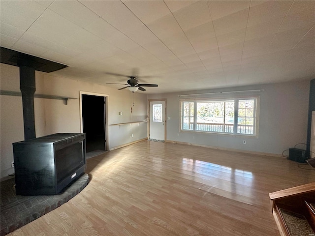 unfurnished living room featuring wood finished floors, a wood stove, a ceiling fan, and baseboards