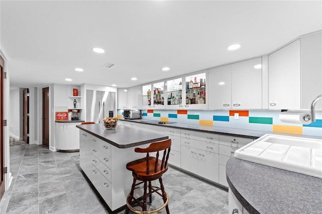 kitchen featuring stainless steel refrigerator with ice dispenser, dark countertops, a kitchen island, white cabinetry, and light marble finish flooring
