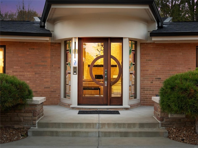 exterior entry at dusk featuring brick siding and a shingled roof